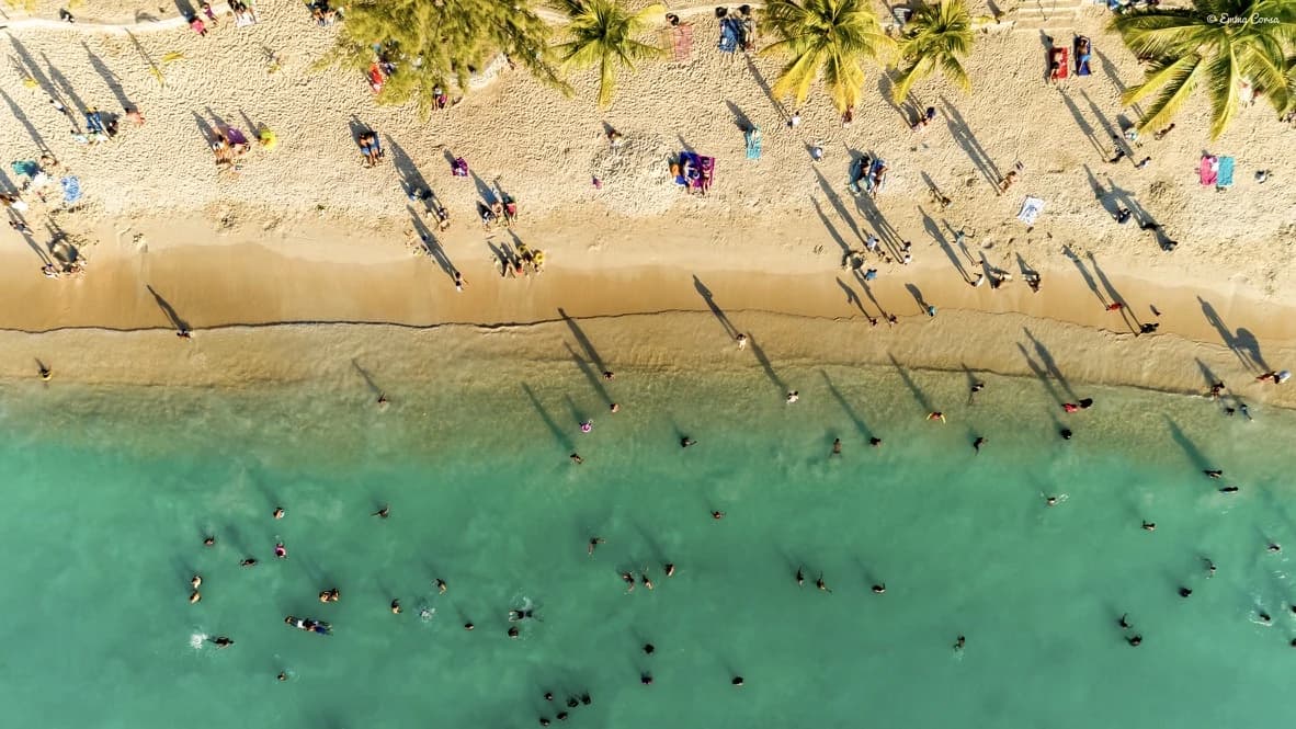 Aerial view of swimmers and long shadows along a turquoise shoreline, with the original Emma Corsa watermark